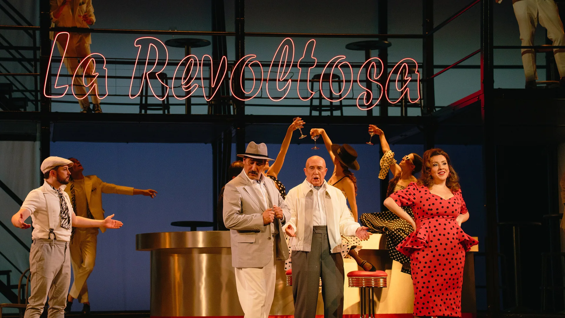 Singers in 1950s-style costume stand on a stage set with a pink neon sign reading La Revoltosa and a bar behind them, in a zarzuela production at a Madrid theatre.