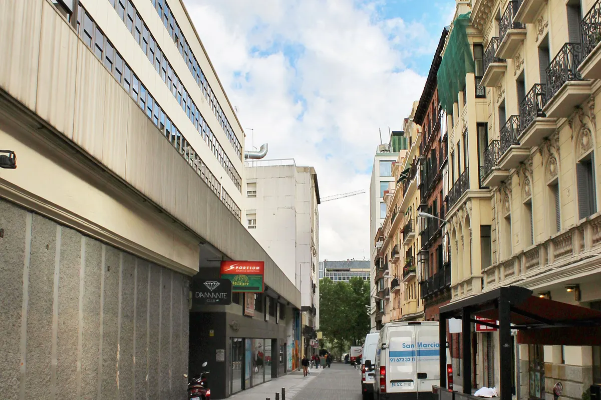 A quiet city street with a long modern building on the left, cream-coloured balconied apartment blocks on the right and a cloudy sky
